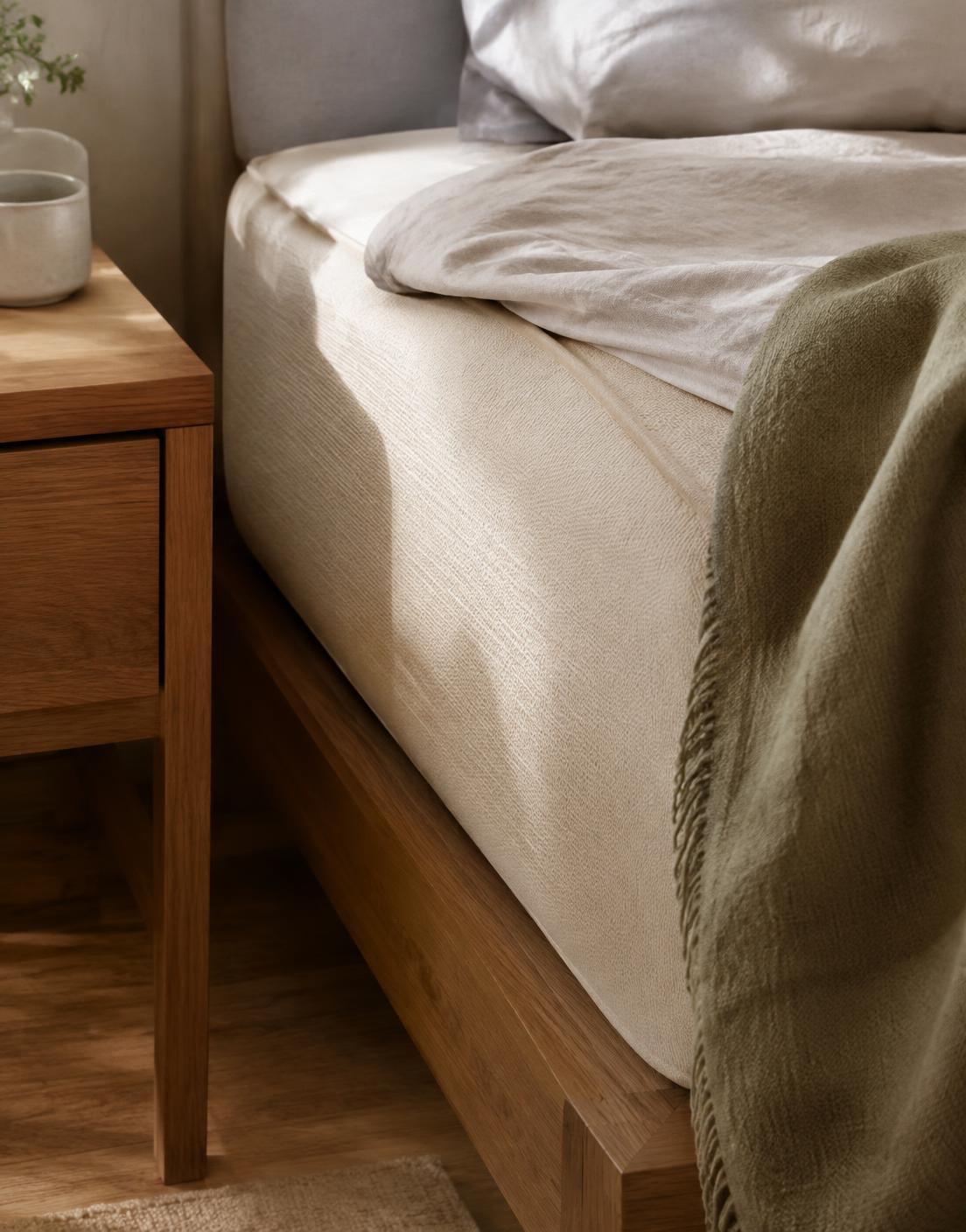 Corner of a natural latex mattress on an oak bed frame beside a timber bedside table, morning light